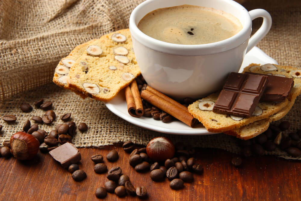 Small cup of espresso, surrounded by biscotti, cinnamon sticks, and coffee beans